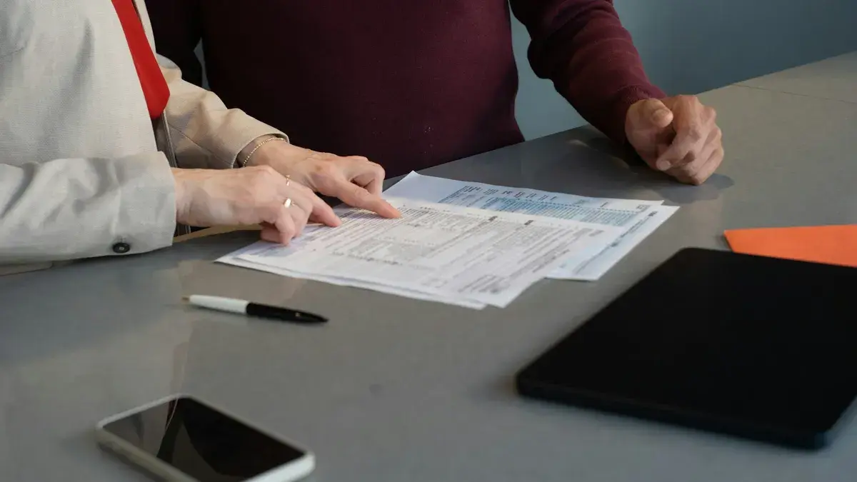 Two people at a table pointing at printed forms, with a pen, phone and tablet beside them