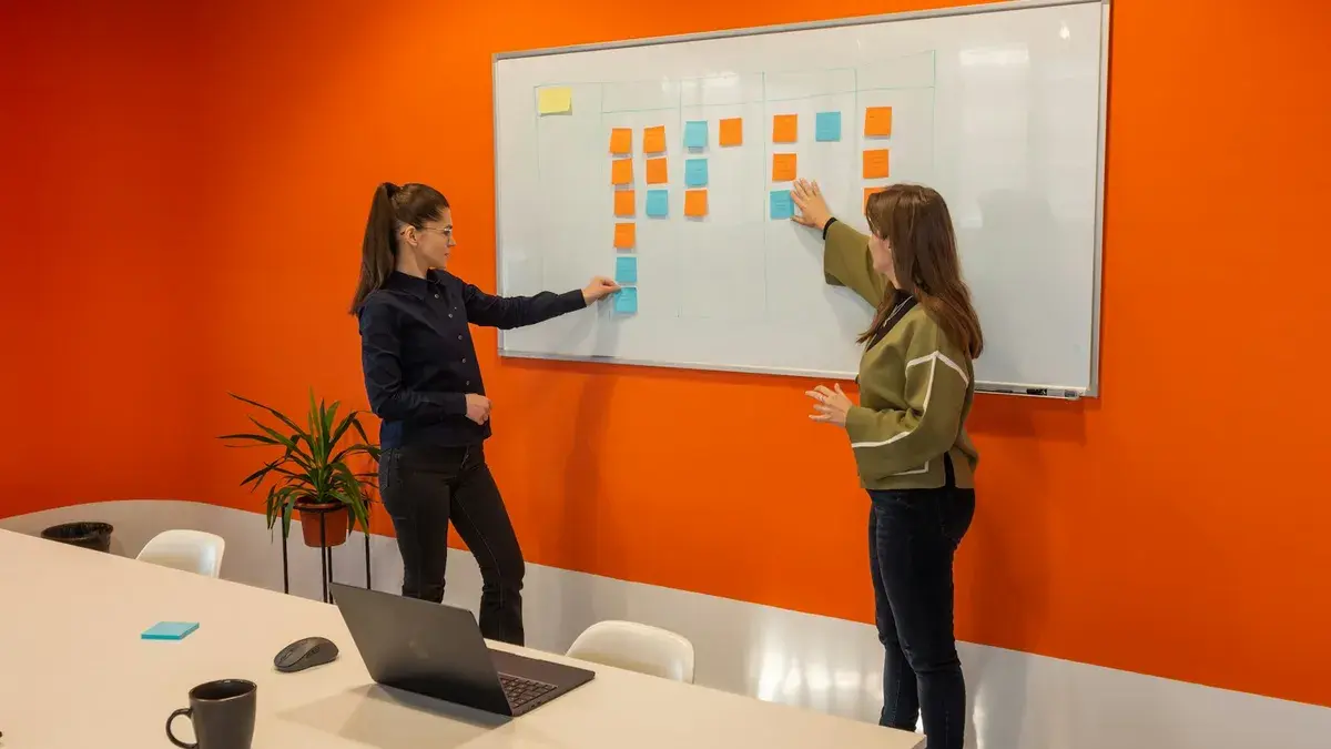 Two colleagues sorting sticky notes into columns on a whiteboard in a meeting room