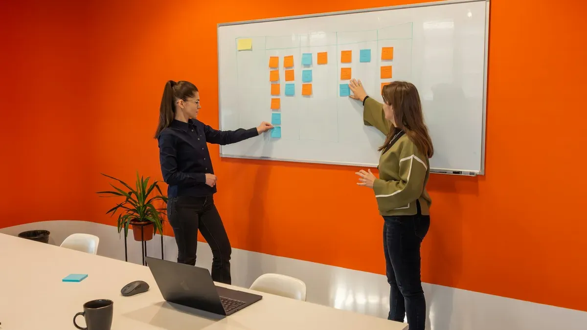 Two colleagues sorting sticky notes into columns on a whiteboard in a meeting room