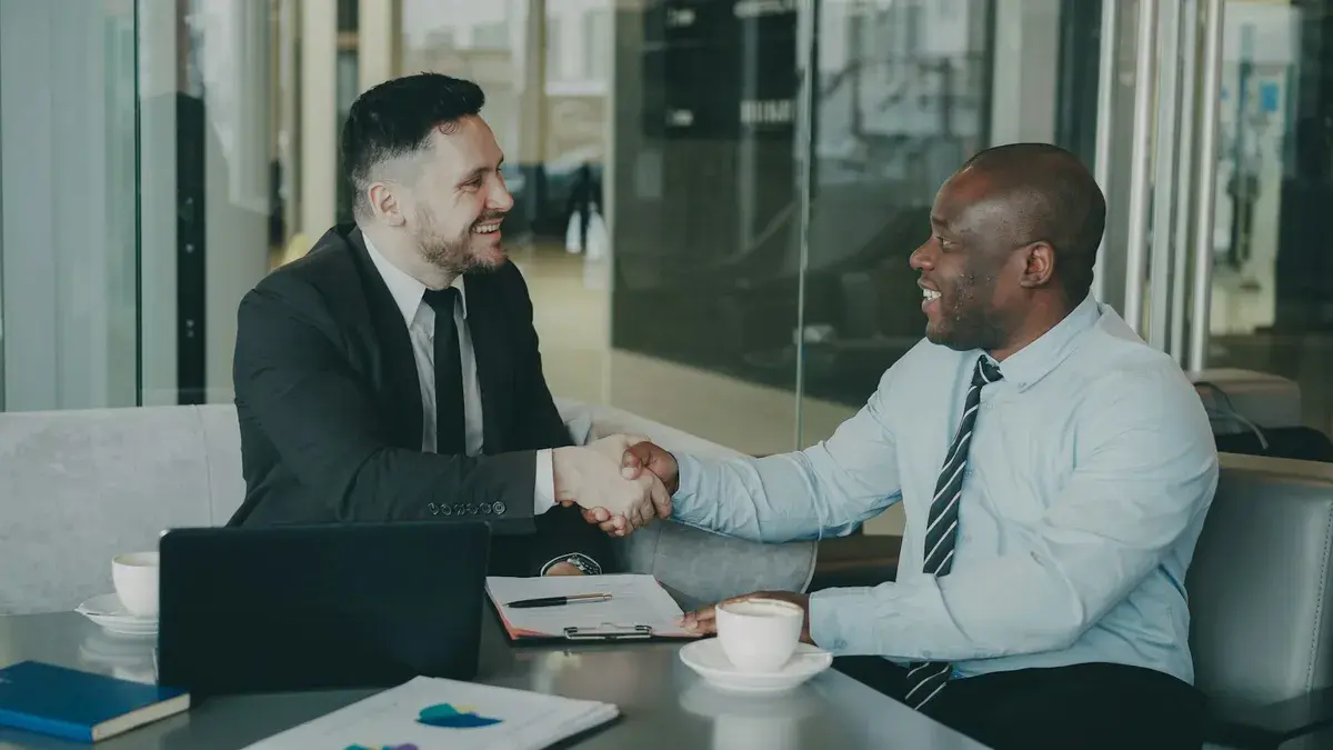 Two businessmen shaking hands across a table in a glass-walled office