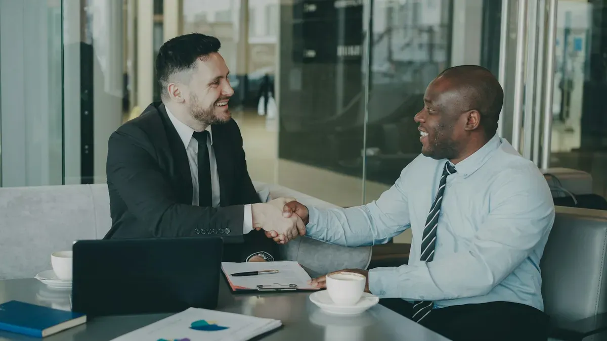 Two businessmen shaking hands across a table in a glass-walled office