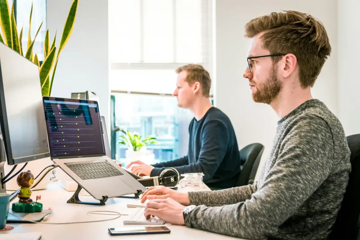 Two IT professionals working at desks in a bright office, one monitoring output on a laptop