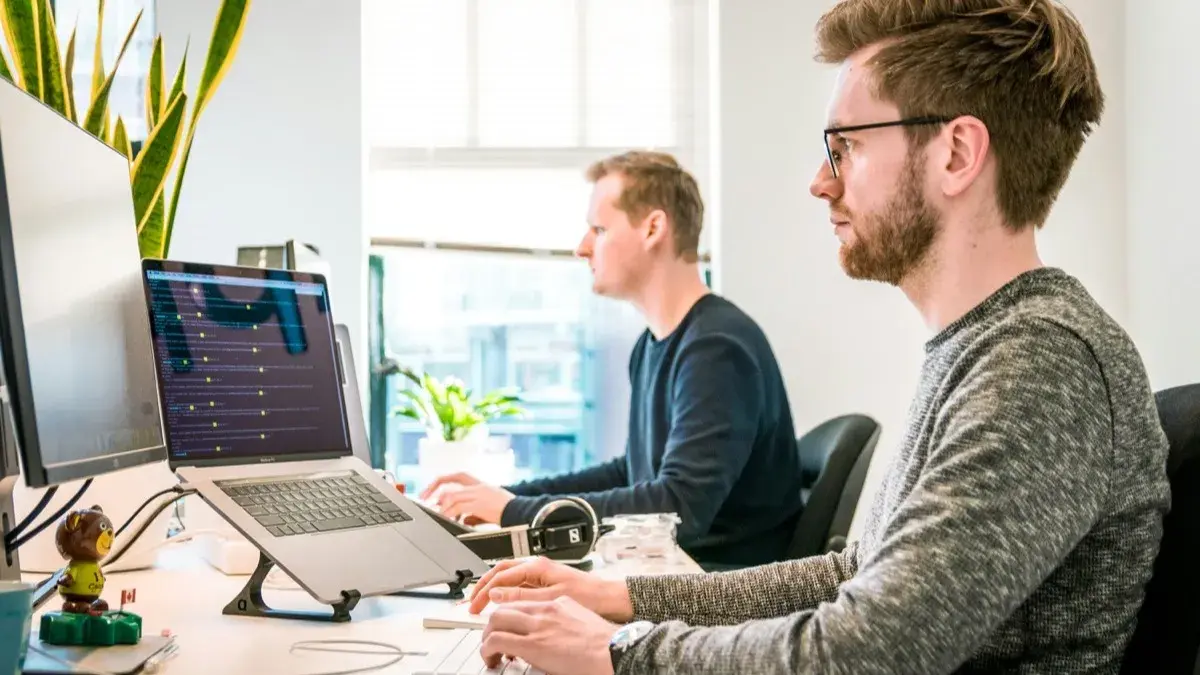 Two IT professionals working at desks in a bright office, one at a laptop with a headset within reach